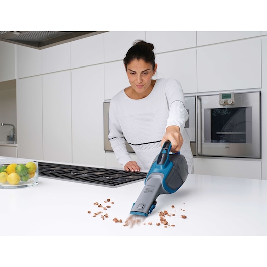 A person uses a BLACK+DECKER handheld vacuum cleaner to clean crumbs from a white kitchen countertop. The kitchen is modern, with built-in ovens and a bowl of fruit visible on the side.