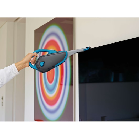 A person uses a BLACK+DECKER handheld vacuum cleaner with an extended nozzle to clean the edge of a television screen. A colorful circular artwork is visible in the background.