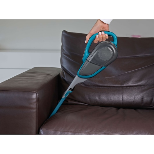 A hand holding a BLACK+DECKER handheld vacuum cleaner model DVJ320J, cleaning the crevice between the seat and arm of a dark brown leather sofa.