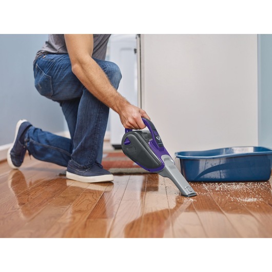 A person is using a BLACK+DECKER handheld vacuum cleaner to clean spilled litter from a hardwood floor next to a blue litter box.
