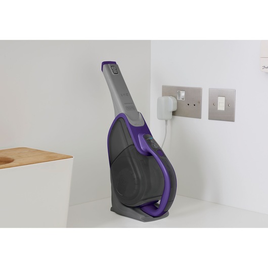 A BLACK+DECKER handheld vacuum cleaner in a charging dock, positioned on a white countertop next to a wall with electrical outlets.