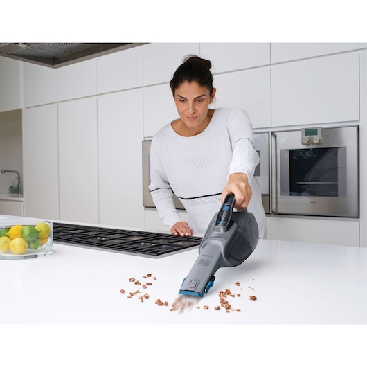 A person using a BLACK+DECKER handheld vacuum cleaner (model DVJ325BF) to clean crumbs from a white kitchen countertop. There are lemons and limes in a glass bowl on the counter, and modern appliances are visible in the background.