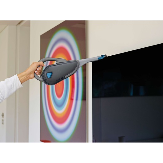 A person is using a BLACK+DECKER handheld vacuum cleaner with a long nozzle to clean the edge of a flat screen TV. In the background, there is a colorful circular artwork on the wall.