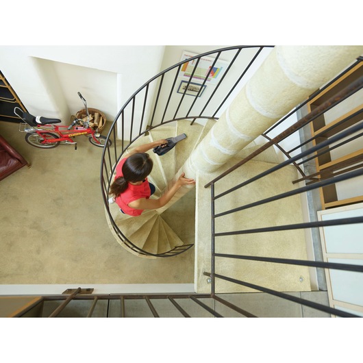 A person is using a BLACK+DECKER handheld vacuum cleaner to clean a spiral staircase in a home. A red bicycle and a basket can be seen on the floor below.