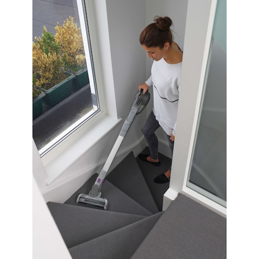 A person cleaning a carpeted staircase with a BLACK+DECKER cordless vacuum cleaner. The vacuum is being used on stairs in a modern, well-lit home with large windows.