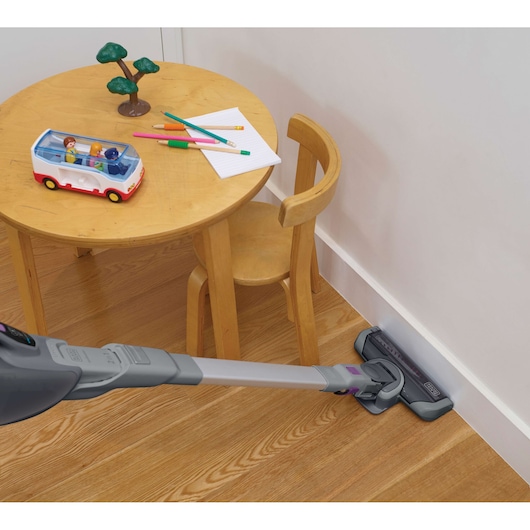 A BLACK+DECKER cordless vacuum cleaner is being used to clean wooden flooring near a small round table and chair. The table has children's toys, colored pencils, and paper on it.