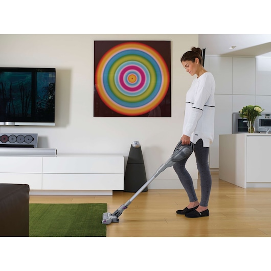 A person using a BLACK+DECKER cordless stick vacuum cleaner on a wooden floor in a modern living room. The person's face is not identifiable. There is a colorful circular artwork on the wall, a television, and a green rug in the room.