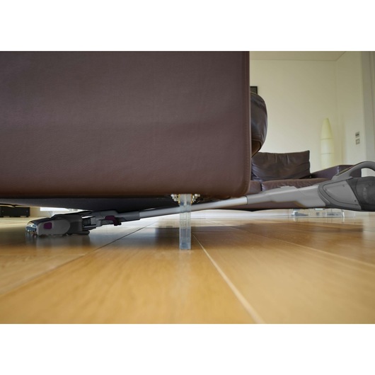 A cordless stick vacuum cleaner being used to clean under a brown sofa with clear legs on a wooden floor in a modern living room.