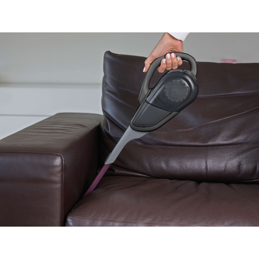 A person using a BLACK+DECKER handheld vacuum cleaner with a narrow nozzle attachment to clean in between the cushions of a dark brown leather sofa.