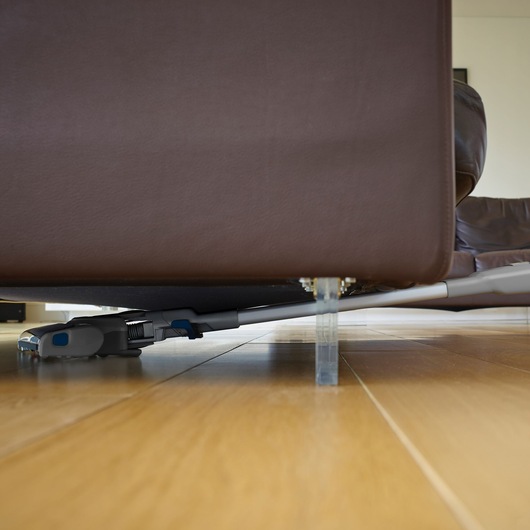 A BLACK+DECKER cordless vacuum cleaner cleaning under a brown sofa on a wooden floor.