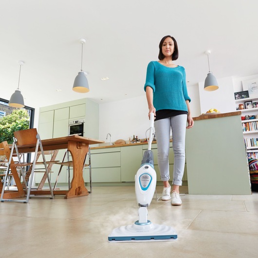 A person with a blurred face uses a BLACK+DECKER steam mop FSM1605 to clean the tiled floor in a modern kitchen. The mop emits steam while being pushed across the floor.