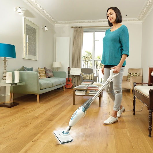 A person is using a BLACK+DECKER FSM1605 steam mop to clean a wooden floor in a modern living room. The room is bright and includes a sofa, guitar, table with books, and large windows with curtains.