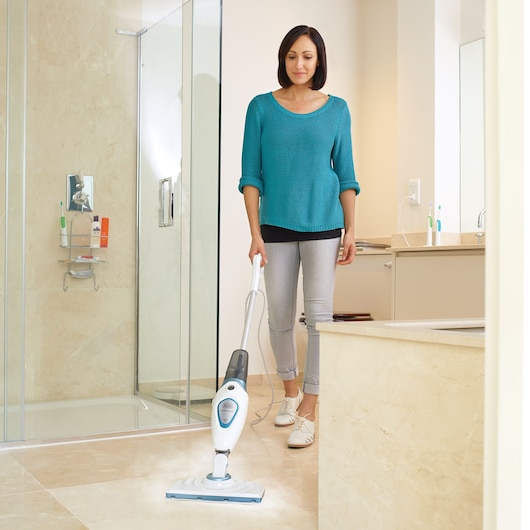 A person cleaning the bathroom floor with a BLACK+DECKER steam mop. The room features a shower, sink, and beige tiled surfaces.