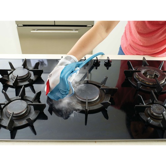 A person uses a steam cleaning glove to clean a black gas stove top. Steam is visible as the glove moves across the stove burner.