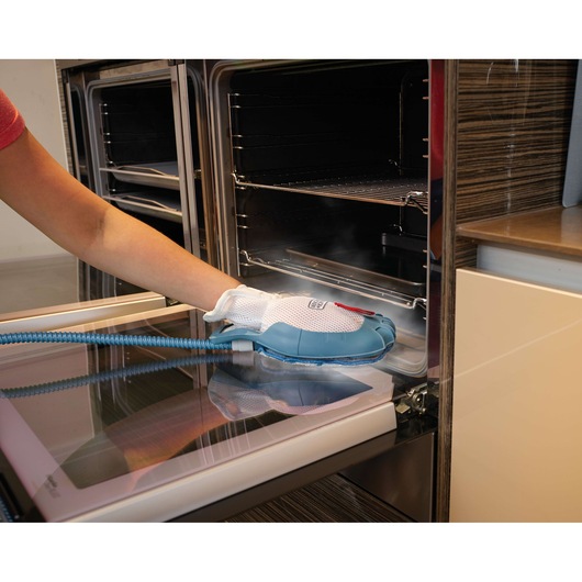 A person uses a BLACK+DECKER steam cleaning glove attachment to clean the inside of an oven, focusing on the oven door.