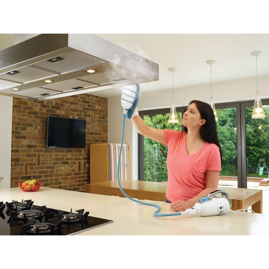 A person using a BLACK+DECKER steam cleaner to clean a kitchen range hood. The steam cleaner produces visible steam while the person holds the cleaning head against the hood. The kitchen features modern lighting, a gas stove, a flat screen, and large windows with a garden view.