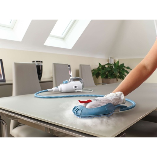 A hand is using a BLUE&DECKER steam cleaning glove to clean a table surface. The steam cleaning device is connected by a blue hose and rests on the table. The scene takes place in a bright room with chairs, a plant, and skylights.