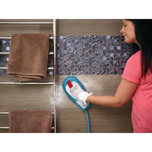 A person wearing a pink shirt uses a BLACK+DECKER steam cleaning mitt to clean wall tiles in a bathroom. A towel rack with folded brown towels is visible next to the tiled wall.