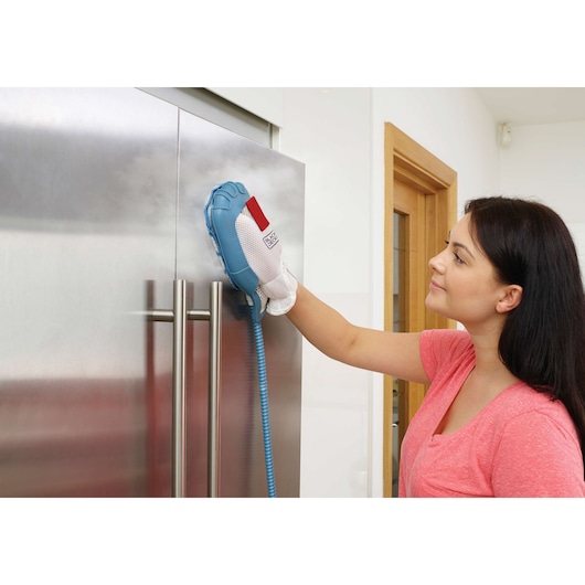 A person uses a BLACK+DECKER steam cleaning glove to clean a stainless steel refrigerator door in a modern kitchen. The person's face is blurred for privacy.