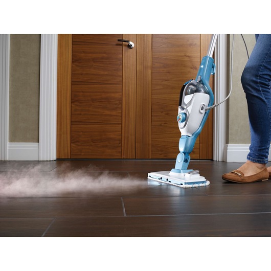 A person using a BLACK+DECKER steam mop to clean a dark tiled floor. Steam is visible coming out from the mop head as it cleans.