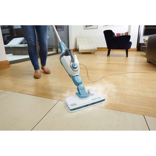 A person cleaning a tiled and wooden floor with a BLACK+DECKER steam mop, model FSMH13101SM, in a modern living room.