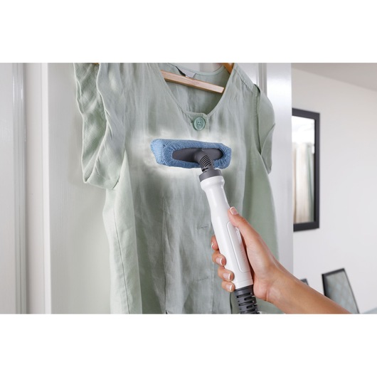 A hand holds a steam cleaning device with a blue attachment, being used to clean or refresh a hanging light green shirt.