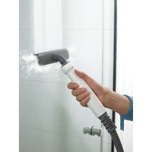 A hand holds a BLACK+DECKER steam cleaning device, using it to clean a glass shower door with visible steam.