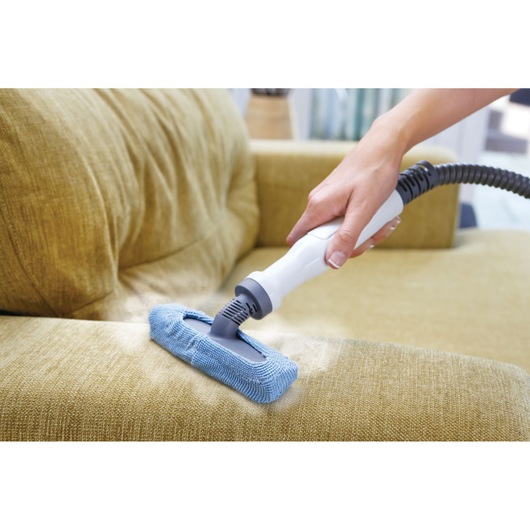 A hand uses a BLACK+DECKER steam cleaning device with a blue microfiber attachment to clean a yellow upholstered sofa.