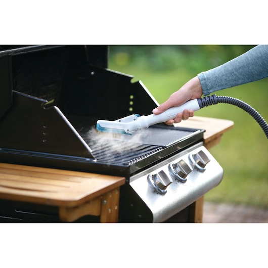 A person is cleaning a barbecue grill with a handheld steam cleaner featuring a hose attachment. Steam is visible coming from the appliance onto the grill grates.