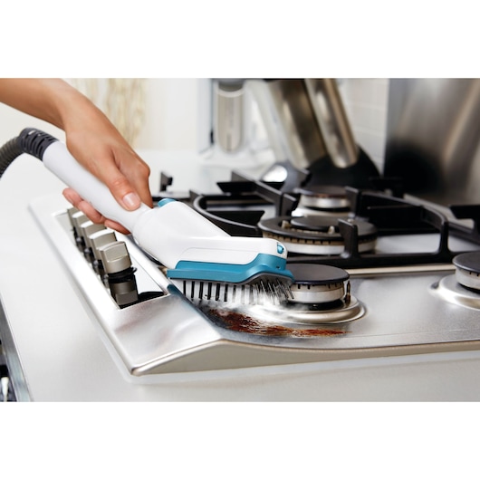 A hand uses a BLACK+DECKER steam cleaner with a brush attachment to clean a stainless steel gas stovetop, removing grime near the burner.