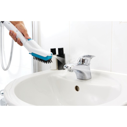 A steam cleaning device being used to clean a bathroom sink faucet. The device has a brush attachment and is emitting steam onto the faucet, with bottles visible on the sink edge.