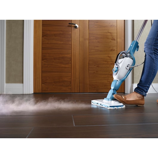 A person is using a BLACK+DECKER steam mop to clean a dark tile floor in front of wooden doors. Steam is visible coming from the mop.