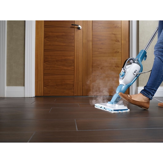 A person uses a BLACK+DECKER steam mop to clean a dark brown tiled floor in front of wooden doors. Steam is visibly coming from the mop head.