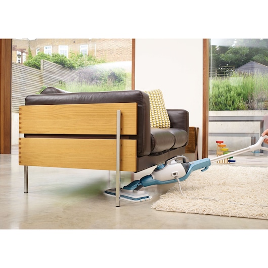 A steam mop cleaning under a modern wooden and leather sofa, with part of a rug visible on a polished floor in a bright room with large windows.
