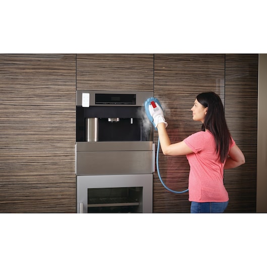 A person using a BLACK+DECKER steam cleaning glove to clean the exterior of a built-in kitchen appliance against a wooden cabinet wall. Steam is visible coming from the glove.