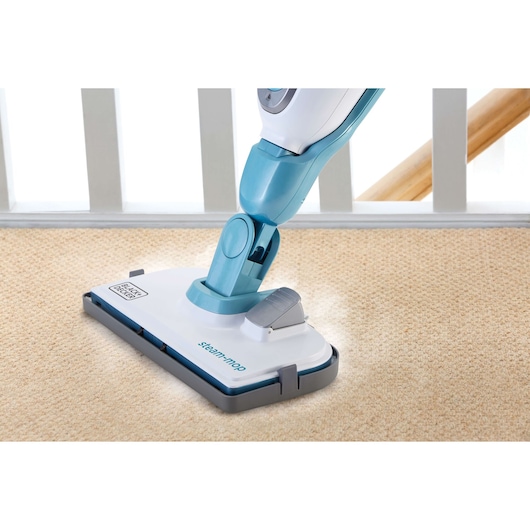 A close-up of a BLACK+DECKER steam mop cleaning a light-colored carpet. The mop head is shown in action, emitting steam, with a railing and wooden banister visible in the background.