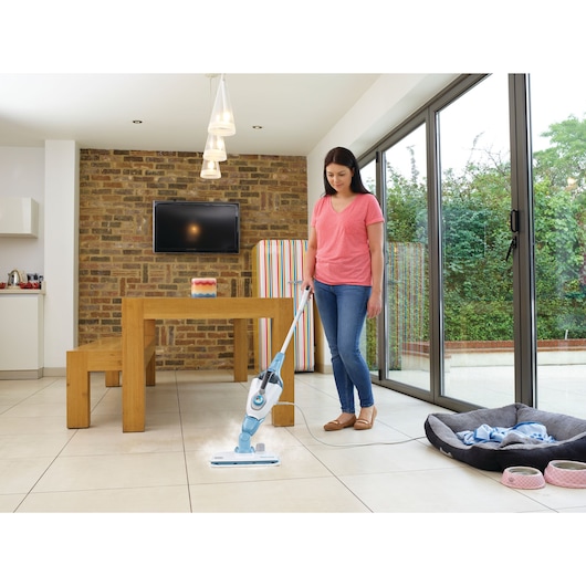 A person is using a BLACK+DECKER steam mop (FSMH13151SM) to clean the tiled floor in a modern dining room with a wooden table and benches, a TV mounted on a brick wall, and large glass doors looking out onto a garden. There is a pet bed and bowls nearby.