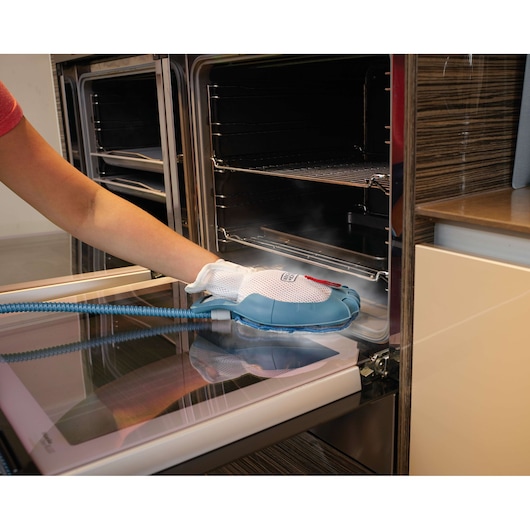 A person is cleaning the inside of an oven using a steam-cleaning hand tool with a glove attached. The tool has a flexible hose and emits steam.