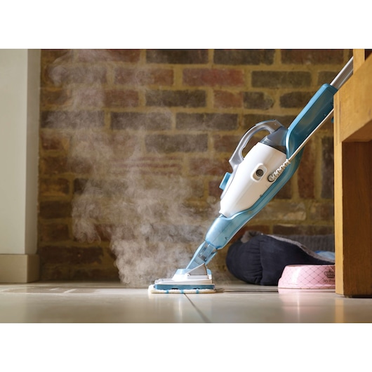 A close-up view of a steam mop cleaning a tiled floor, with visible steam rising from the mop head. The mop is blue and white and is positioned next to a wooden furniture piece and pet accessories in a room with a brick wall.