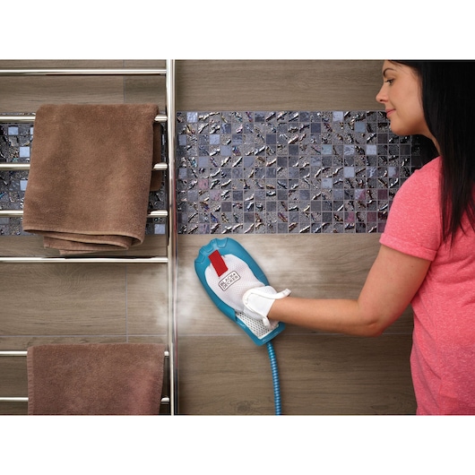 A person uses a BLACK+DECKER steam cleaning glove on tile wall in a bathroom. There are brown towels hanging on a metal rack next to the wall.