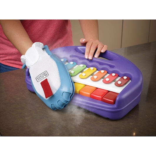 A person uses a BLACK+DECKER steam cleaning glove to clean a colorful children's xylophone toy placed on a countertop. Steam is visible coming from the glove.