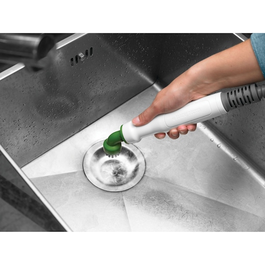 A hand using a BLACK+DECKER portable cleaning tool with a green brush attachment to clean a stainless steel sink.