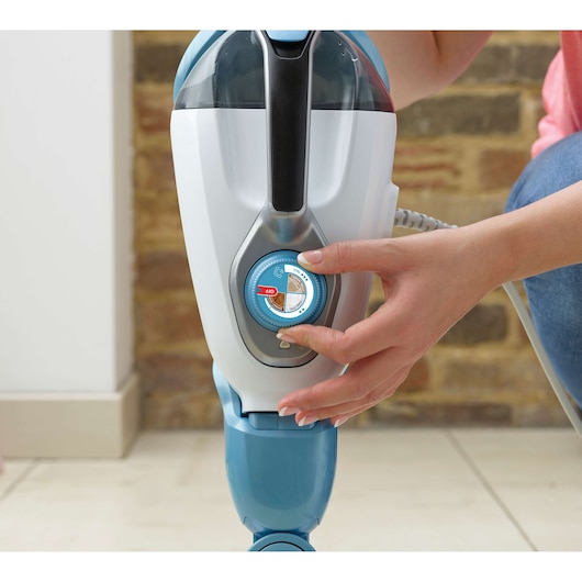 A close-up view of a person adjusting the control dial on a BLACK+DECKER steam mop. The steam mop is white and blue, with a clear water tank and a labeled dial for settings.