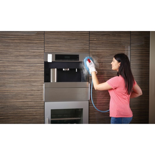 A person using a BLACK+DECKER steam cleaning glove to clean a built-in kitchen appliance with wood paneling in the background. Steam is visible coming from the glove.
