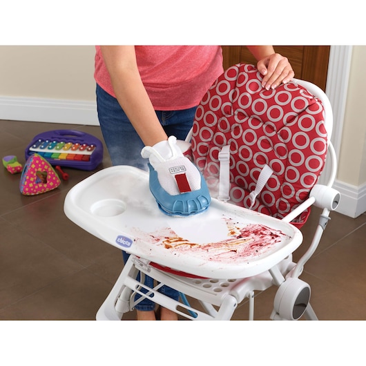 A person is using a BLACK+DECKER steam cleaning glove to clean food stains from a high chair tray. The high chair has a red and white patterned seat, and children's toys are visible on the floor in the background.