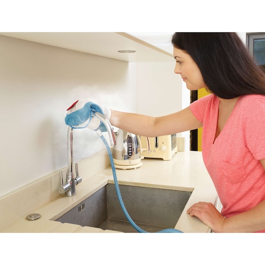 A person using a BLACK+DECKER steam cleaner to clean a kitchen faucet and surrounding area. The person is wearing a pink shirt and their face is blurred. Kitchen appliances like a kettle and toaster are visible on the countertop.
