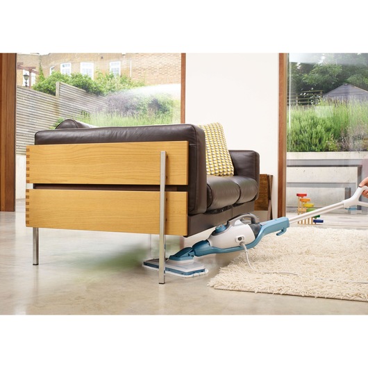 A steam mop cleaning the floor under a modern wooden and leather sofa in a bright living room, next to a white rug and large windows showing a garden outside.