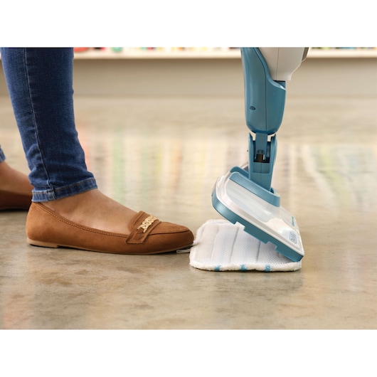 A close-up of a person wearing brown loafers and blue jeans, using their foot to attach a cleaning pad to a blue and white BLACK+DECKER steam mop on a polished floor.