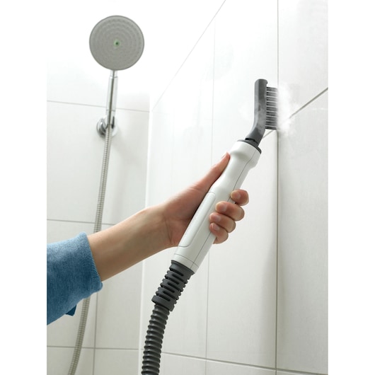 A person uses a BLACK+DECKER steam cleaning tool with a brush attachment to clean the grout between white shower tiles.