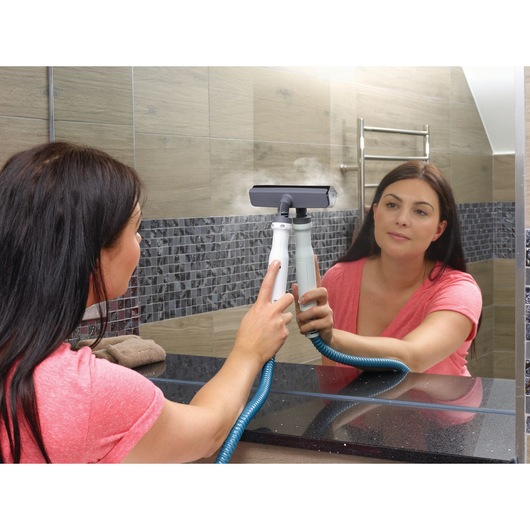 A person using a BLACK+DECKER steam cleaner with a hose attachment to clean a bathroom mirror, with steam visible on the glass.